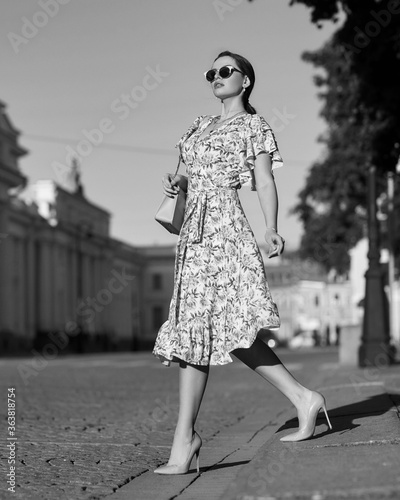 Pretty lady wearing wrap around dress. Beautiful woman with wavy brunette hair walking un the city. Elegant girl with long leegs in high heels. Fashionable female model in sunglasses holding handbag
