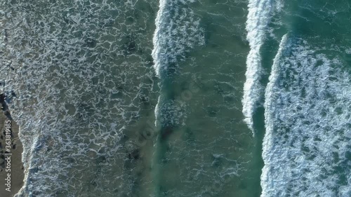 Aerial flying forward downward view of ocean waves crashing onto beach with rocks adding texture to sand through water.