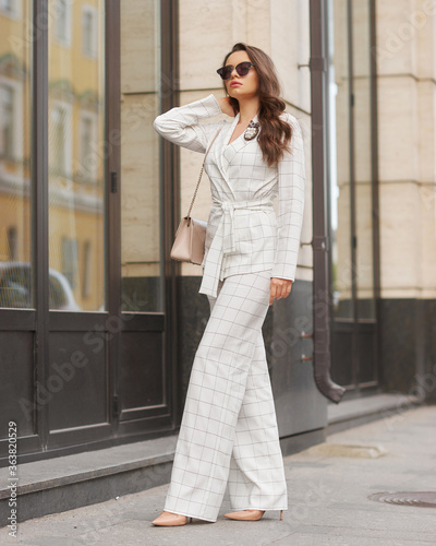 Young beautiful elegant woman in white casual costume walking at street on a summer day. Pretty lady wearing sunglasses and holding handbag. Businesswoman standing and posing in the city