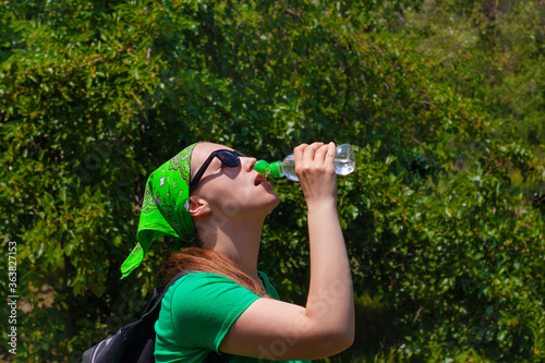 Young girl holds a bottle of water in hand and drinks. Portrait of pretty hipster in sunglasses, green shirt, bandana stands on the nature on sunny day. Weight loss, summer active lifestyle body care.
