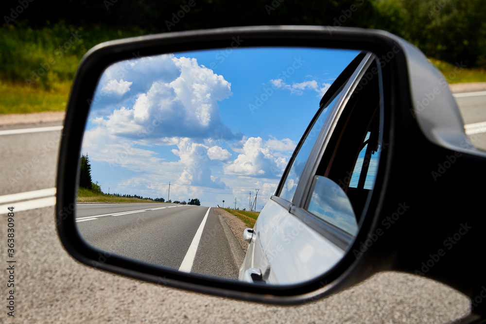 Side mirror of the car with reflection of white clouds in the blue sky and the road in the background.