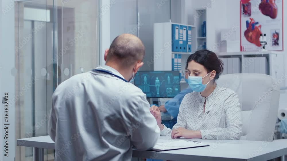 Woman at doctor appointment during COVID-19 wearing a mask and gloves, talking with medic through a protective wall. Medical consultation in protective equipment concept shot of sars-cov-2 global Woman at doctor appointment during COVID-19 wearing a mask and gloves, talking with medic through a protective wall. Medical consultation in protective equipment concept shot of sars-cov-2 global