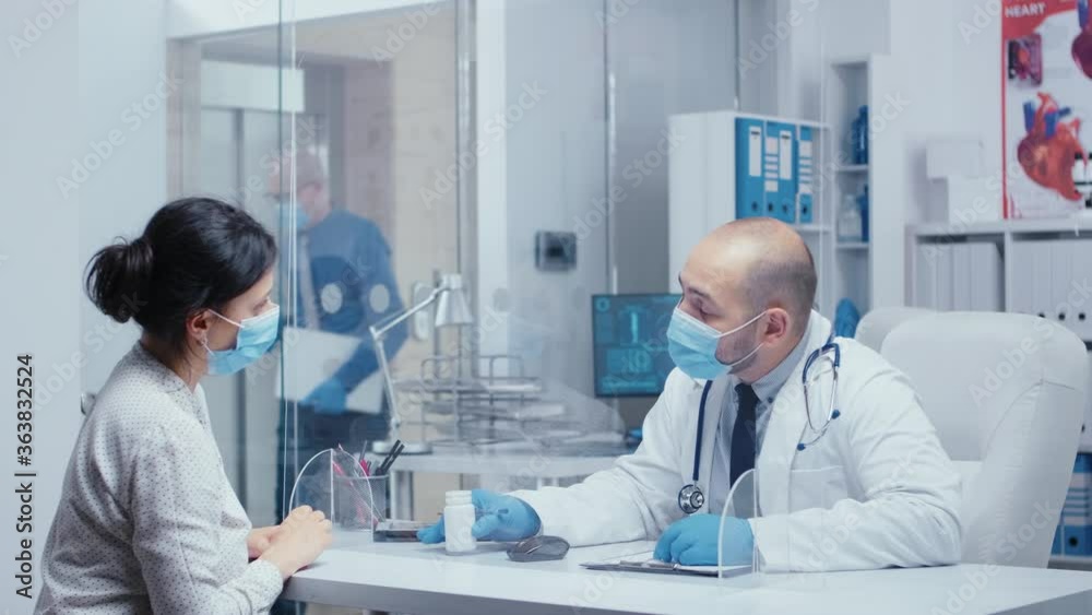 Male doctor prescribing drugs to woman patient during COVID-19 global health crisis, they are talking through a plexiglass wall, wearing masks and gloves. Medical consultation in protective equipment Male doctor prescribing drugs to woman patient during COVID-19 global health crisis, they are talking through a plexiglass wall, wearing masks and gloves. Medical consultation in protective equipment