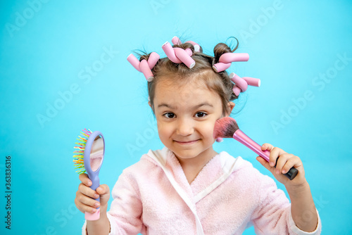A smiling little girl with curlers in her hair is looking at the camera on a blue isolated background. A little girl in curlers holds a comb in her hand mirror and a brush for blush. Empty space for t