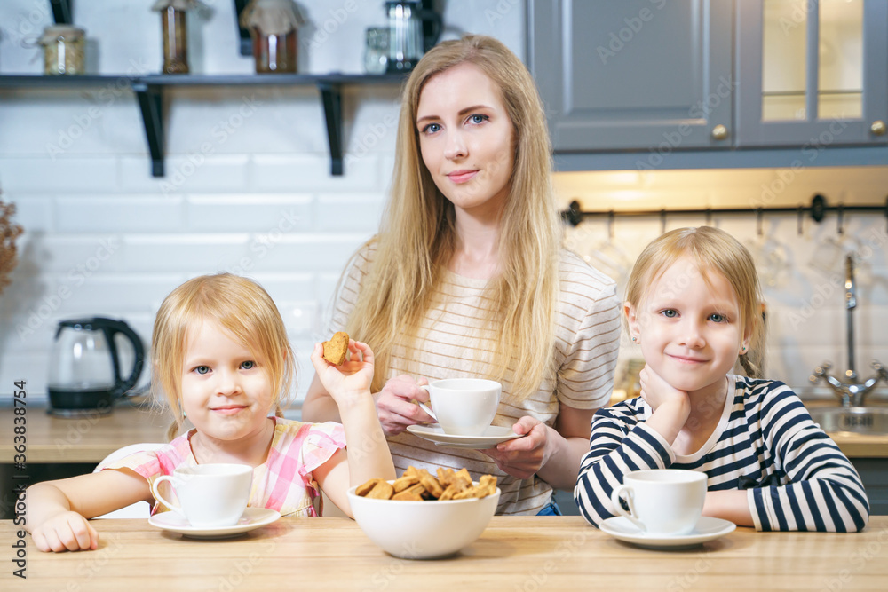 Portrait of a happy family young mother and two daughters drinking tea or milk with freshly baked homemade cookies in the kitchen