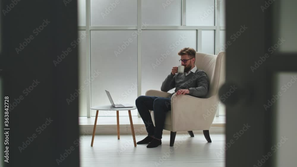 Side View of Young Businessman in Glasses Sitting on Armchair in Spacious Office Centre Rest Zone, Working, Typing on Laptop, Drinking Coffee or Tea. Side View of Young Businessman in Glasses Sitting on Armchair in Spacious Office Centre Rest Zone, Working, Typing on Laptop, Drinking Coffee or Tea.