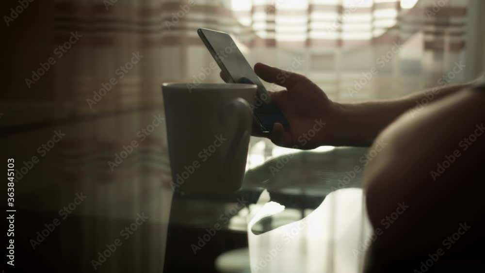 Man with a mobile phone is sitting at a table with a mug of hot coffee, on a light background of the window Man with a mobile phone is sitting at a table with a mug of hot coffee, on a light background of the window