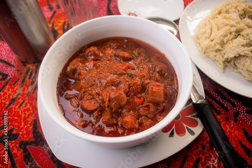 A delicious white bowl of red meat sauce or beef gravy filled with diced pieces of meat, tomatoes, onions and pepper served on a colorful black and red pattern table cloth with some mashed pasta