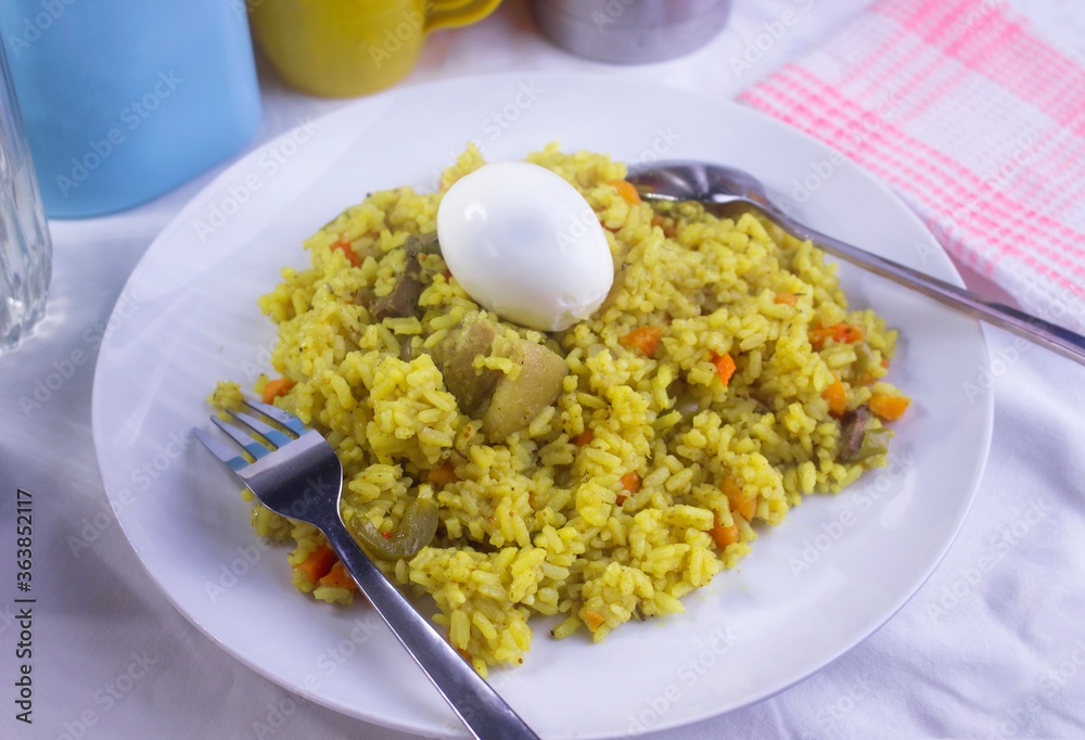 A delicious meal of Nigerian fried rice cooked with some chopped meat, diced carrots and a boiled egg on top. A fork and spoon are placed at the sides of the white plate on a white table cloth  