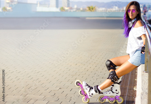Roller skating young Caucasian girl with violet hair, sunglasses and protection outdoors sitting on parapet and relaxing after rollerblading on inline skates. outdoor summer activities. Space for text
