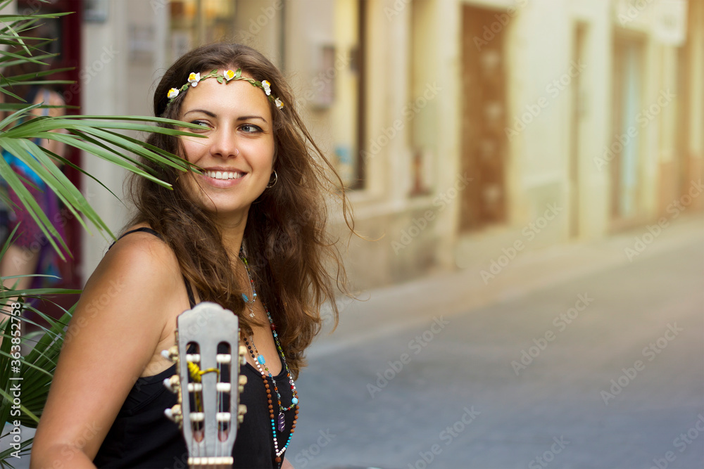 Fototapeta premium beautiful happy young girl with curly hair hippie style with a guitar and a wreath on her head next to a palm branch. Woman photo portrait waist-high on the street. Free lifestile. Street musicians