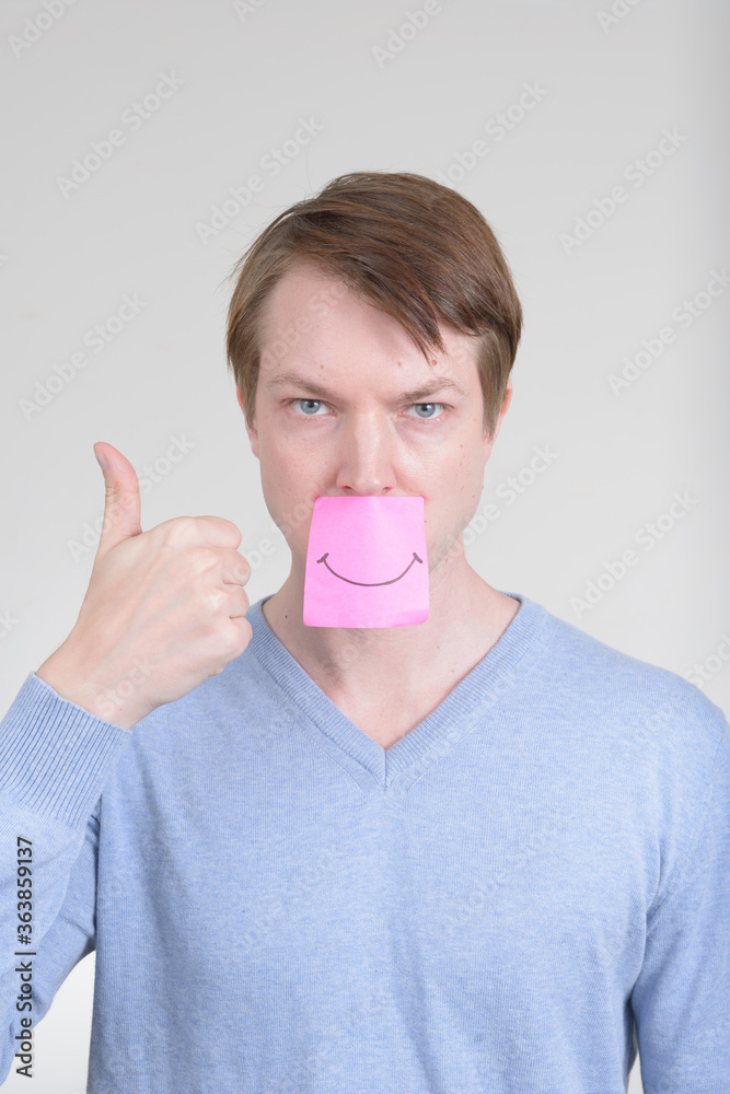 Portrait of young handsome man with smiling sticker covering mouth and giving thumbs up Portrait of young handsome man with smiling sticker covering mouth and giving thumbs up