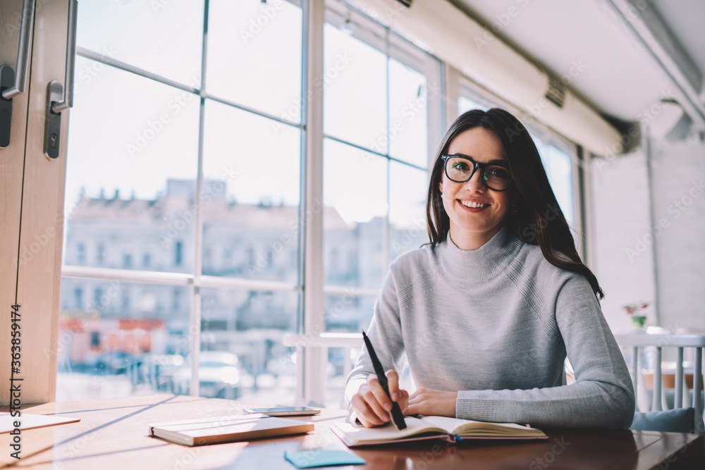 Portrait of happy young trendy woman working as journalist resting at cafeteria, successful hipster girl in stylish eyeglasses smiling at camera and creating notes for homework while sitting at cafe