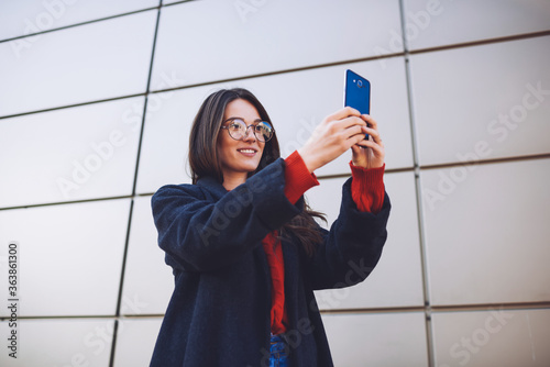 Good looking cheerful woman dressed stylish overcoat making picture using application on modern telephone connected to wireless internet and sharing image for friends in social network while strolling