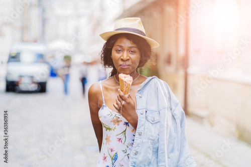 Cheerful black girl in stylish hat with dessert while walking outdoor, selective focus, shallow depth of field. Young smiling African female is eating ice cream in street at summer time.