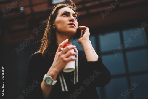 Young attractive woman having mobile conversation on cellular connected to wireless internet during summer strolling around city.Gorgeous teenager walking outdoors with take away coffee and gadget