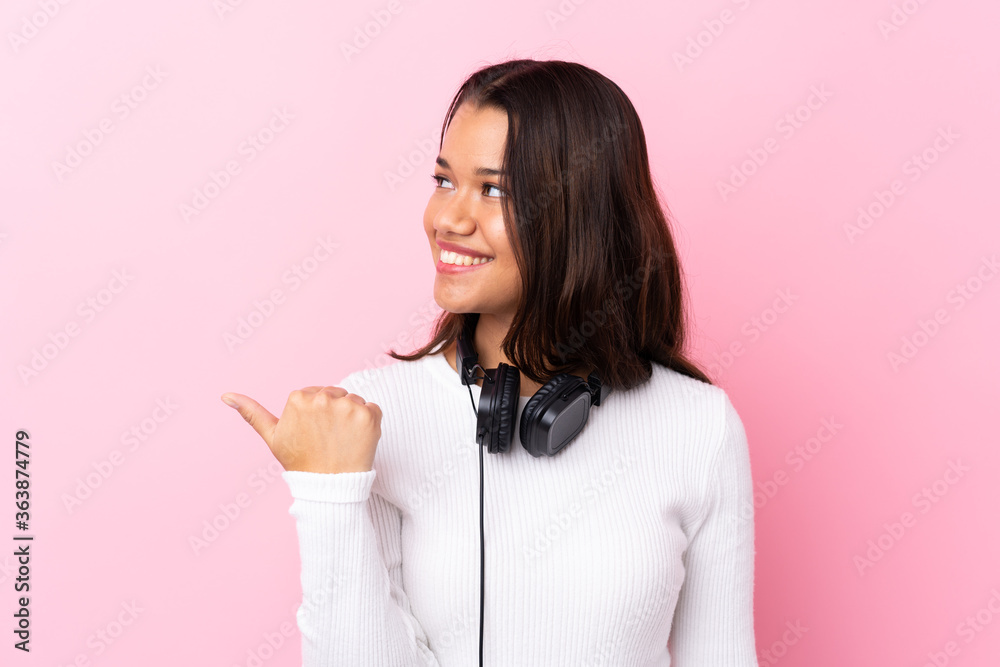 Young woman with earphones over isolated pink wall pointing to the side to present a product Young woman with earphones over isolated pink wall pointing to the side to present a product
