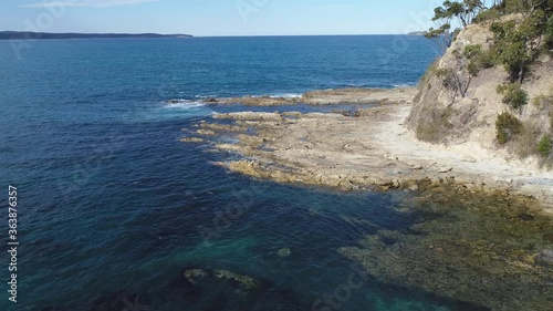 Aerial fly over azure blue ocean with reef and rocks near a small coastal point.