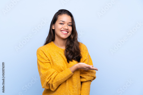 Young Colombian girl with sweater over isolated blue background presenting an idea while looking smiling towards