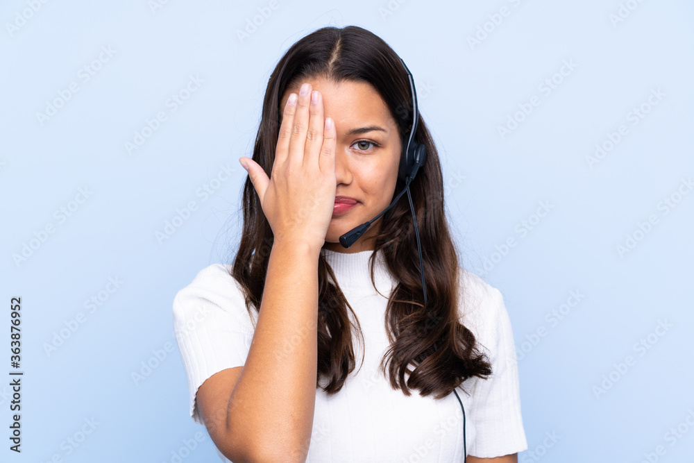 Young telemarketer Colombian woman over isolated blue background covering a eye by hand Young telemarketer Colombian woman over isolated blue background covering a eye by hand