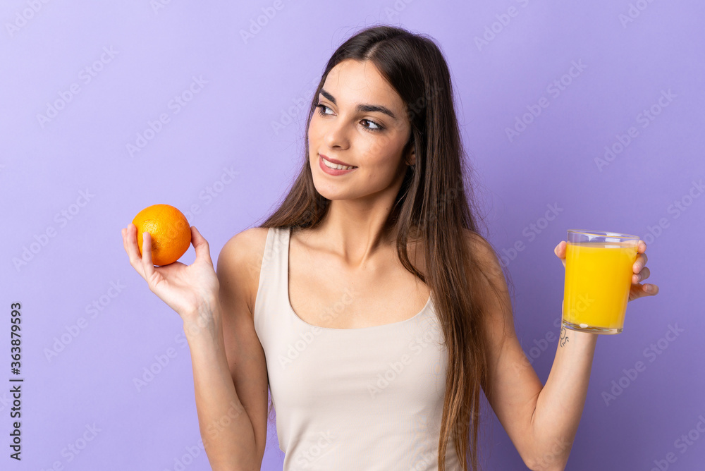 Young caucasian woman isolated on purple background holding an orange and an orange juice Young caucasian woman isolated on purple background holding an orange and an orange juice