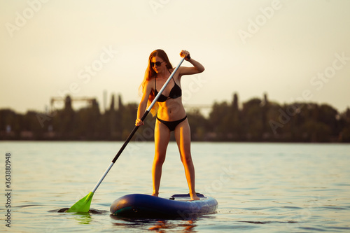 Warm light. Young attractive woman standing on paddle board, SUP. Active life, sport, leisure activity concept. Caucasian woman on travel board in summers evening time. Vacation, resort, enjoyment.