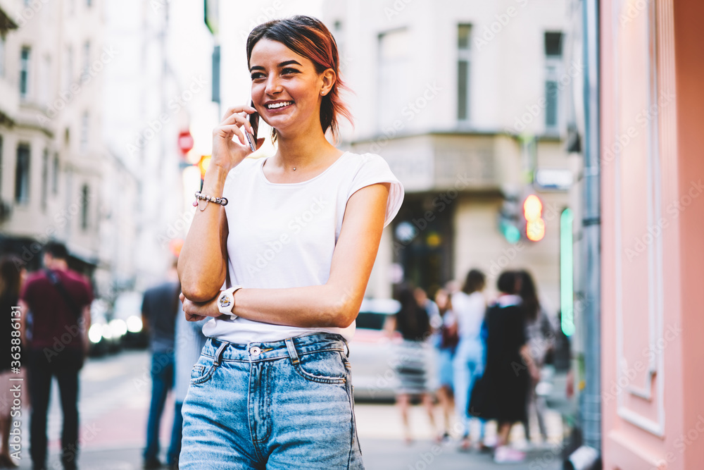 Attractive successful female person dressed in stylish outfit talking on mobile phone with friends while walking at urban setting.Cheerful hipster girl enjoying leisure time at street with gadget