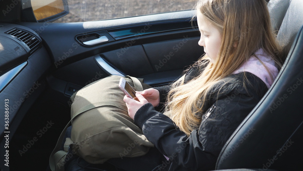 Happy little girl with long blonde hair sits in front passenger seat of modern SUV and uses her smart phone. Beautiful female child browses social media on mobile phone, turns to camera and smiles