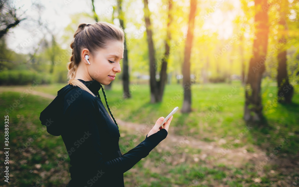 Young smiling girl making sport and running in the park using her phone to listen the music with wireless headphones on sunset in the city watching the screen Young smiling girl making sport and running in the park using her phone to listen the music with wireless headphones on sunset in the city watching the screen