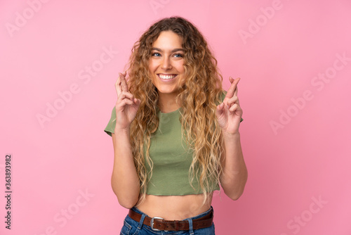 Young blonde woman with curly hair isolated on pink background with fingers crossing