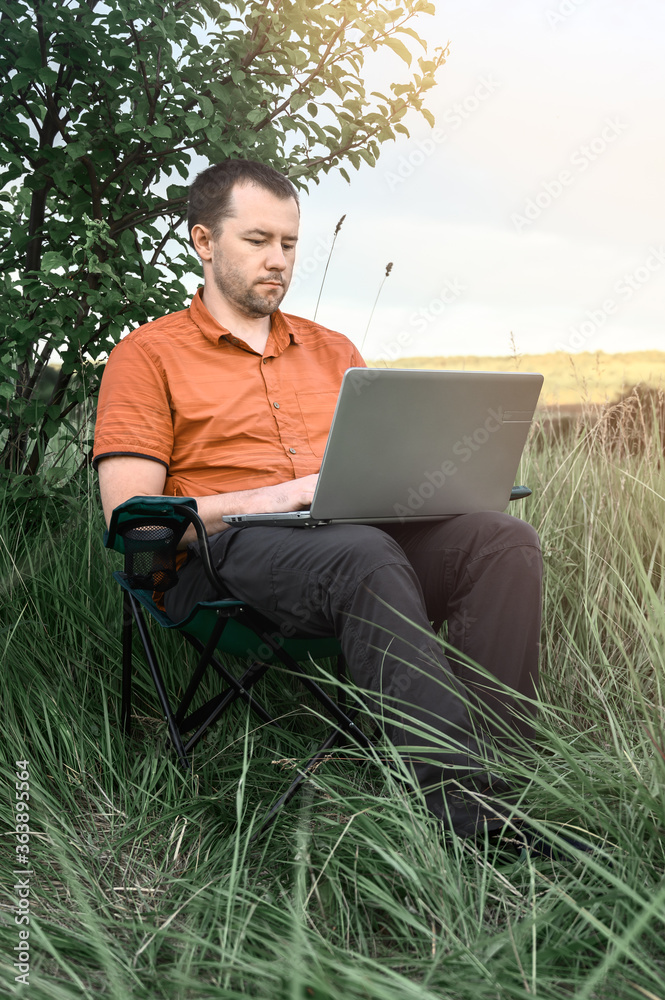 Young caucasian man in orange shirt working on his laptop sitting on a camping chair in meadow. Copy space. Young caucasian man in orange shirt working on his laptop sitting on a camping chair in meadow. Copy space.