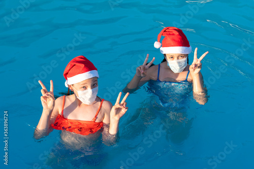 Two little girls in santa claus hat and medical mask swim in the pool. New Year 2021 during the pandemic. The concept of the second wave of coronavirus in the world