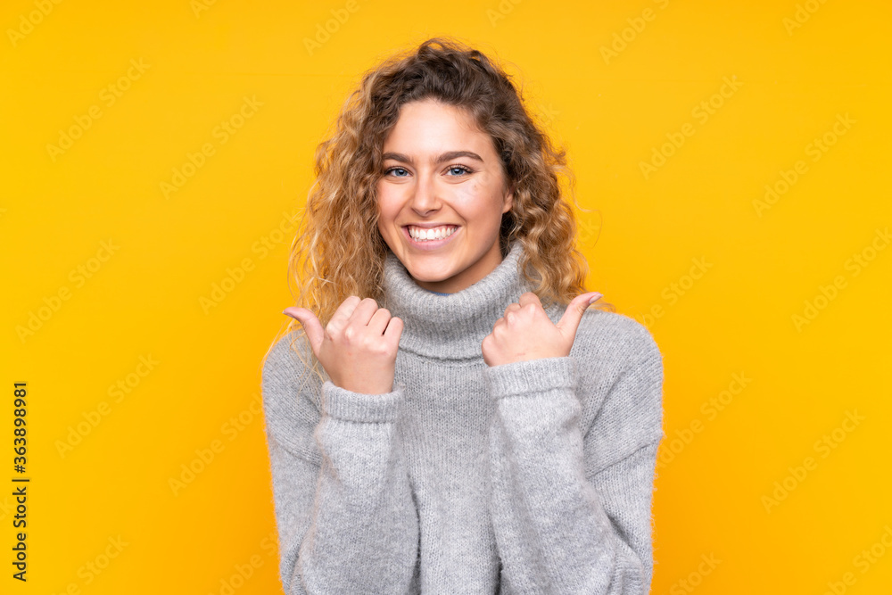 Young blonde woman with curly hair wearing a turtleneck sweater isolated on yellow background with thumbs up gesture and smiling