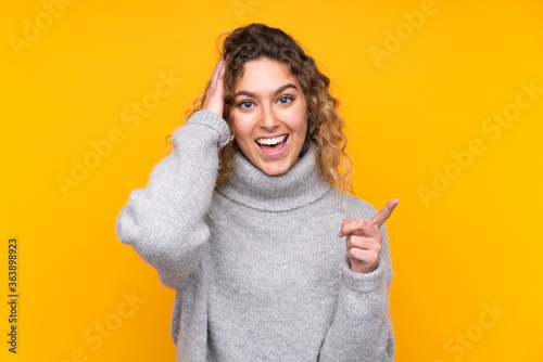 Young blonde woman with curly hair wearing a turtleneck sweater isolated on yellow background surprised and pointing finger to the side © luismolinero