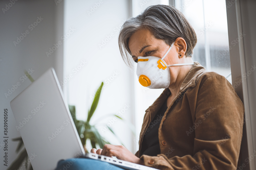 Pensive gray-haired woman in casual clothes and protective mask works on laptop computer sitting on windowsill workplace. Pandemic concept. Side view from below. Tinted image. Pensive gray-haired woman in casual clothes and protective mask works on laptop computer sitting on windowsill workplace. Pandemic concept. Side view from below. Tinted image.