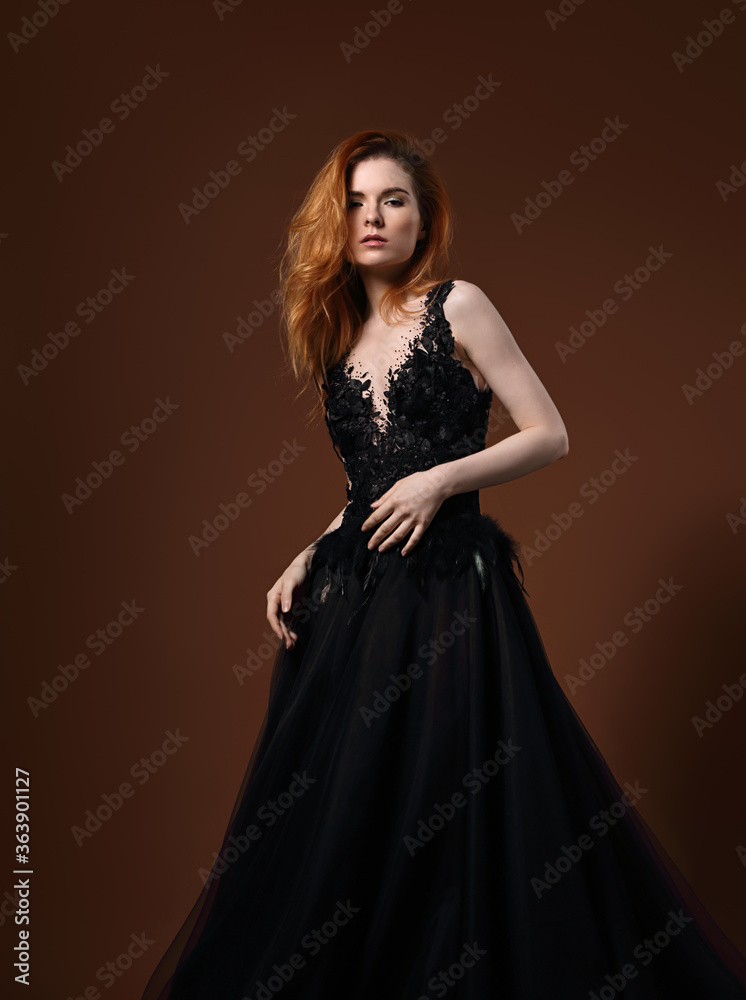 Beautiful elegant young model with bright redhead hairstyle posing in fashion expensive black wedding dress with long skirt on studio bright orange background. Portrait Beautiful elegant young model with bright redhead hairstyle posing in fashion expensive black wedding dress with long skirt on studio bright orange background. Portrait