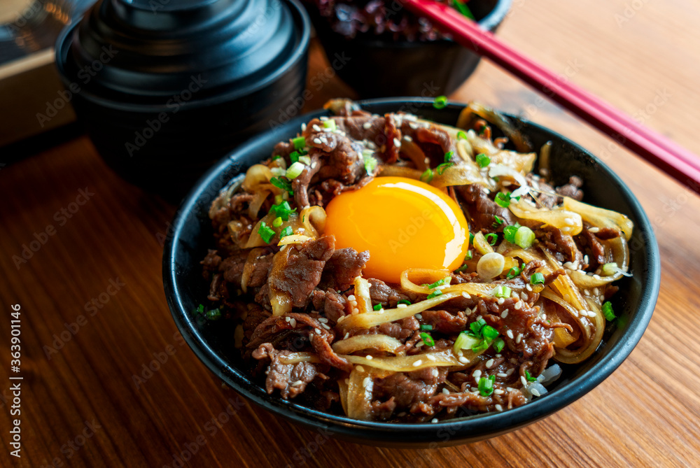 Japanese food, stir fried beef and onion over rice, topping with green onion, white sesame and fresh chicken egg yolk. Food in black bowl on wooden table, natural light, close up food, selected focus. Japanese food, stir fried beef and onion over rice, topping with green onion, white sesame and fresh chicken egg yolk. Food in black bowl on wooden table, natural light, close up food, selected focus.