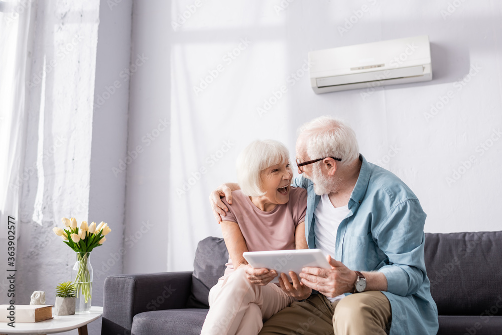 Obraz premium Senior man holding digital tablet and embracing smiling wife on couch