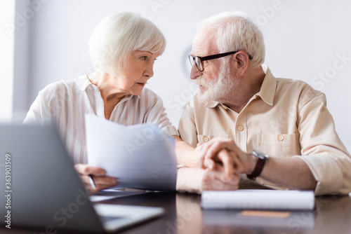 Selective focus of elderly couple holding hands near papers and laptop on table