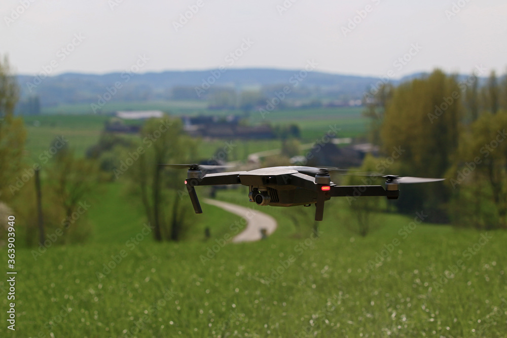 A generic drone hoovering over a beautiful meadow, with a curvy road in a landscape on the background. The camera is under the rotating blades. Flying a drone is the newest hype in aviation technology A generic drone hoovering over a beautiful meadow, with a curvy road in a landscape on the background. The camera is under the rotating blades. Flying a drone is the newest hype in aviation technology