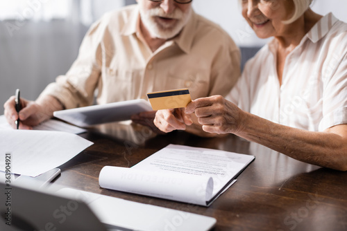 Selective focus of senior woman holding credit card while husband writing on papers near gadgets on table