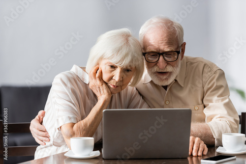 Selective focus of senior man embracing wife while using laptop near cups of coffee and smartphone on table