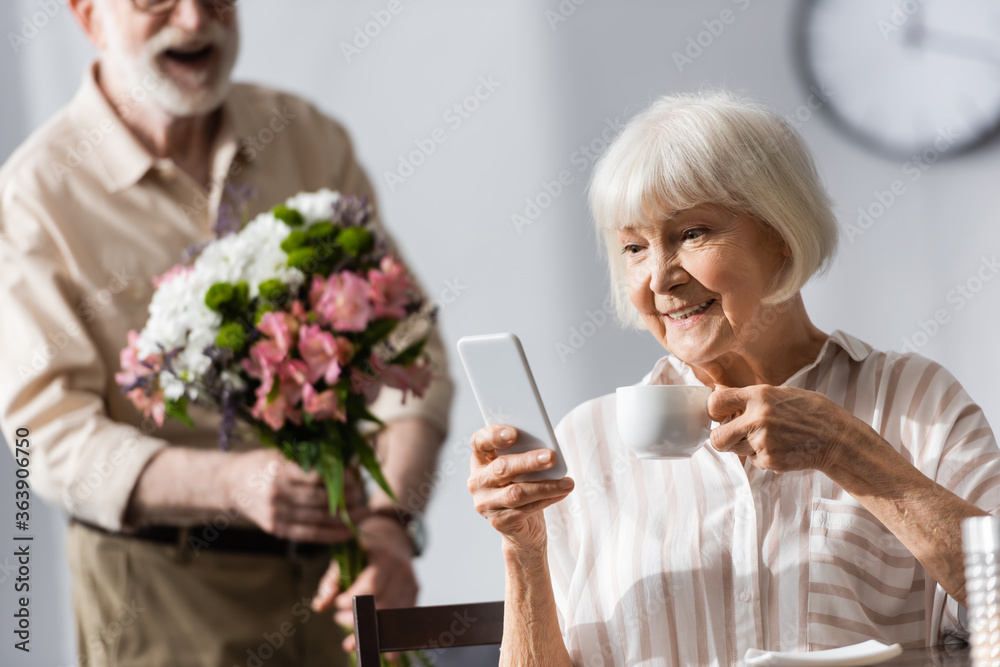 Selective focus of smiling woman holding cup of coffee and using smartphone near husband with bouquet Selective focus of smiling woman holding cup of coffee and using smartphone near husband with bouquet