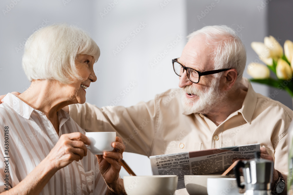 Selective focus of senior man holding newspaper and embracing smiling wife with cup of coffee