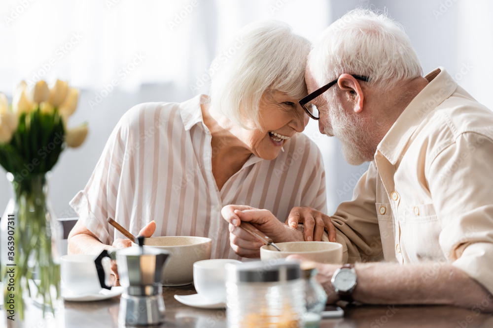 Selective focus of positive senor couple looking at each other near coffee and breakfast on table Selective focus of positive senor couple looking at each other near coffee and breakfast on table
