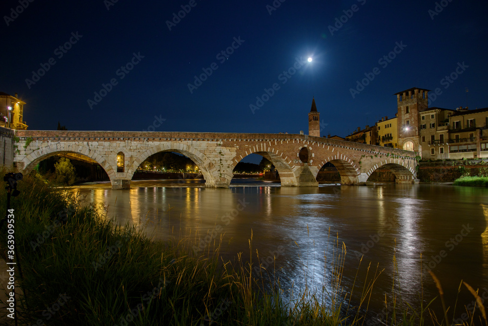 Night photo with moon in sky view along Adige river with view of Ponte della Pietra with tower and bell tower Santa Anastasia, city of Verona, Italy.