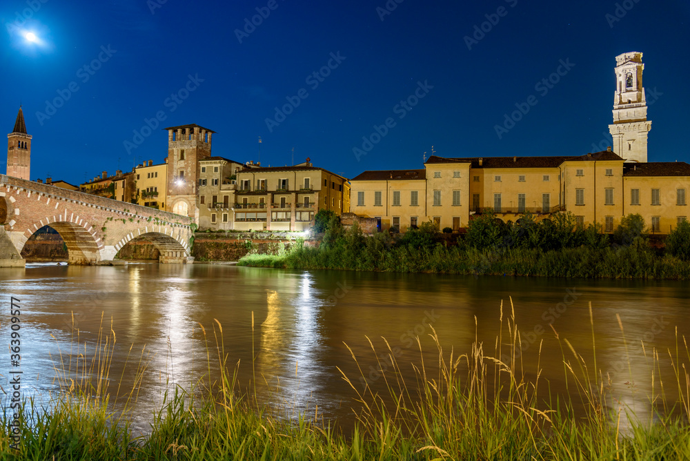 Night photo with moon in sky view along Adige river with view of Ponte della Pietra with tower and bell tower of Santa Anastasia and bell tower of the cathedral, city of Verona, Italy.