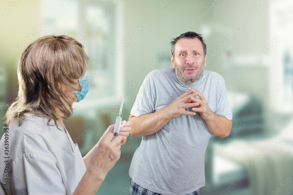 Mature man suffers from an extreme fear of needles. It seems he is going to faint. He is shaking with fear in a hospital. A nurse with a syringe stands nearby. Mature man suffers from an extreme fear of needles. It seems he is going to faint. He is shaking with fear in a hospital. A nurse with a syringe stands nearby.