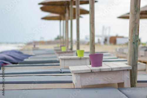 Focus on the cup on the wooden table next to the sun lounger on the beach bar. The beach is empty and there is no one.