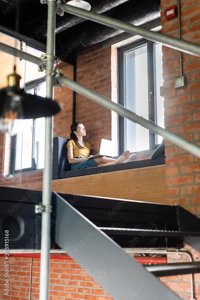 selective focus of businesswoman sitting on window bench with laptop and looking at windows in office